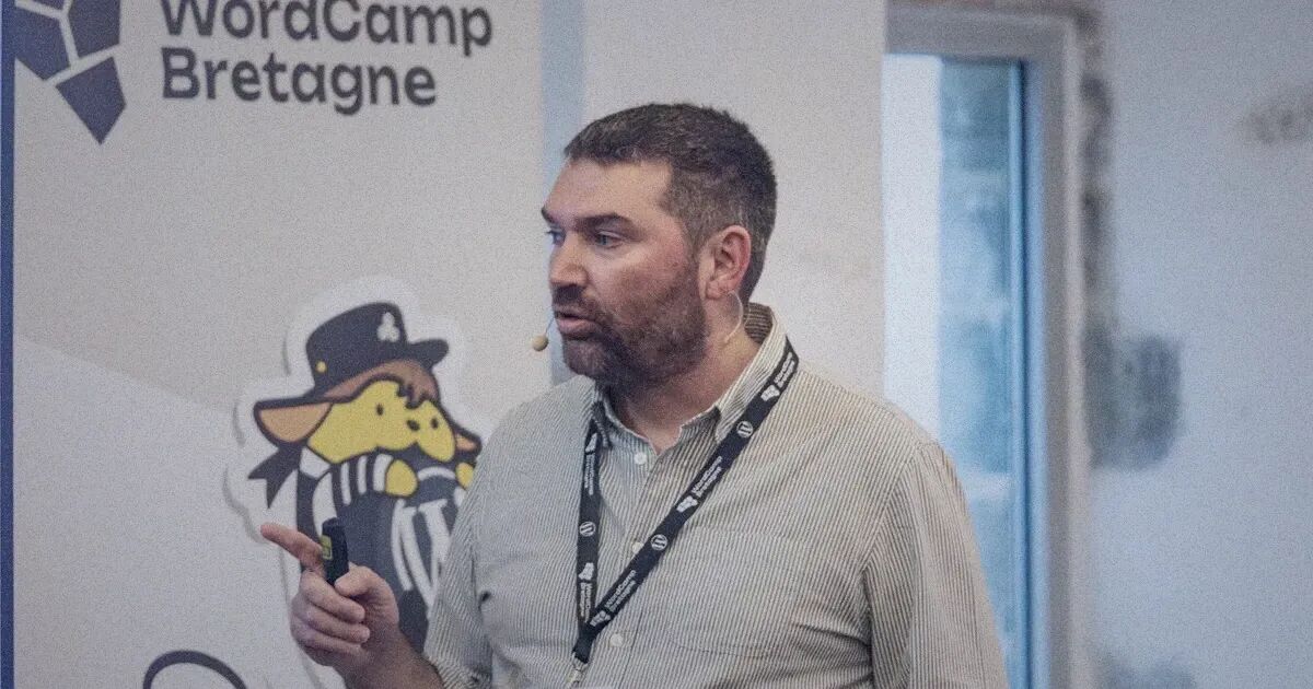 Intervenant micro-casque et badge autour du cou, présentant sa conférence devant le kakémono du WordCamp Bretagne et sa mascotte Wapuu